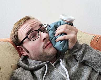 Man holding an ice pack to the side of his jaw while laying on a couch