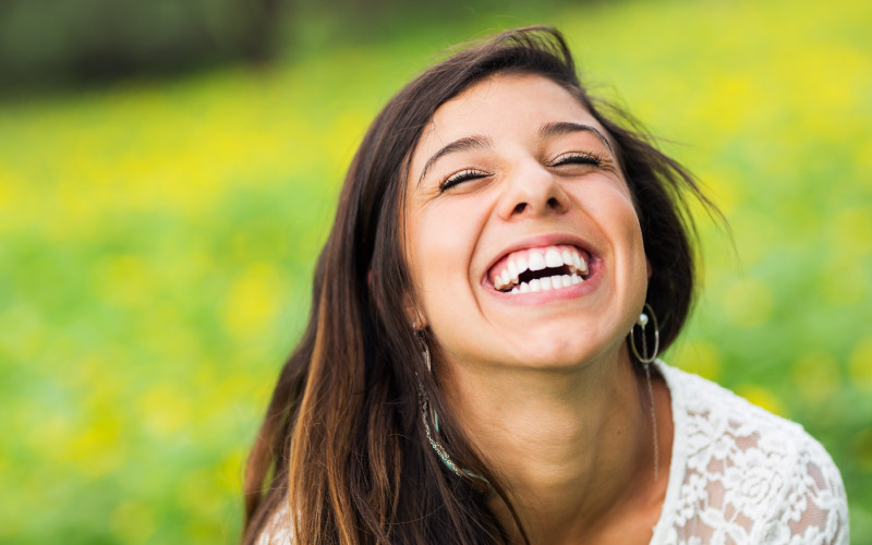 Closeup of a brunette woman smiling with her eyes closed as she laughs in a green and yellow meadow