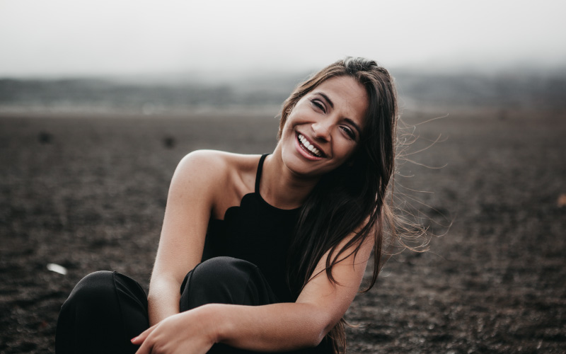 Brunette woman smiles with a bright white smile thanks to dental veneers while sitting elegantly in a dirt field