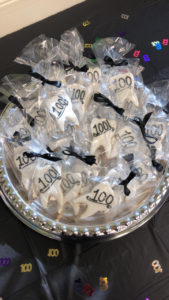 A platter of tooth-shaped cookies with white icing and 100 written on them in baggies with a black ribbon