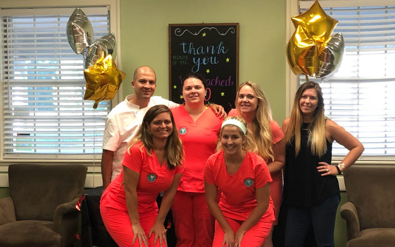 Dr. Quiros and his dental team pose in coral scrubs next to decorations celebrating 100+ Google reviews