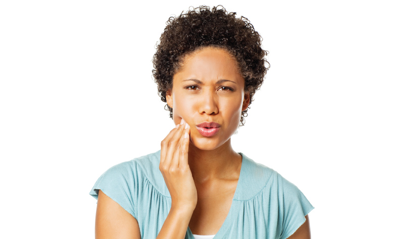 Curly-haired woman wearing a blue blouse touches her cheek in pain due to sensitive teeth